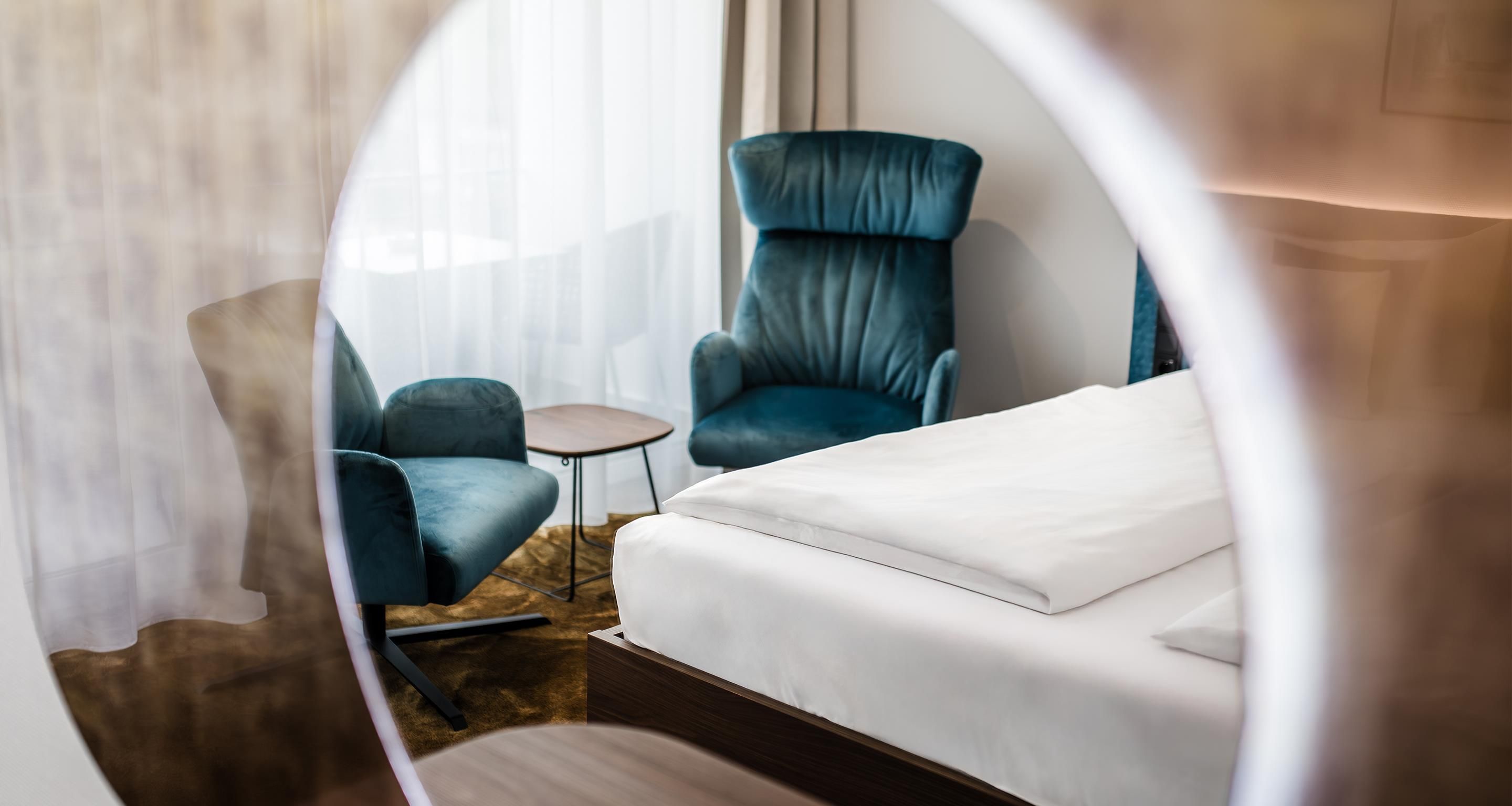 A modern hotel room with a bed, a blue armchair, a small round table, and a view through a circular window.