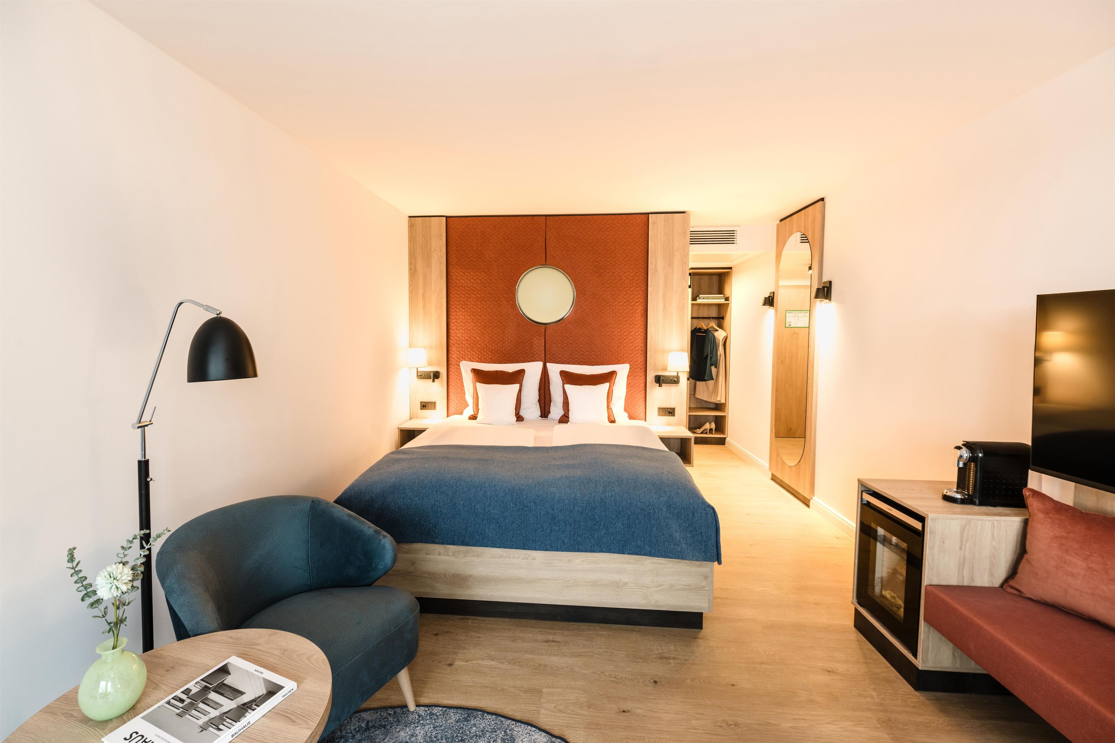Une chambre d’hôtel comprenant un lit, un coin salon avec un fauteuil, une petite table avec un vase, une télévision et une armoire.