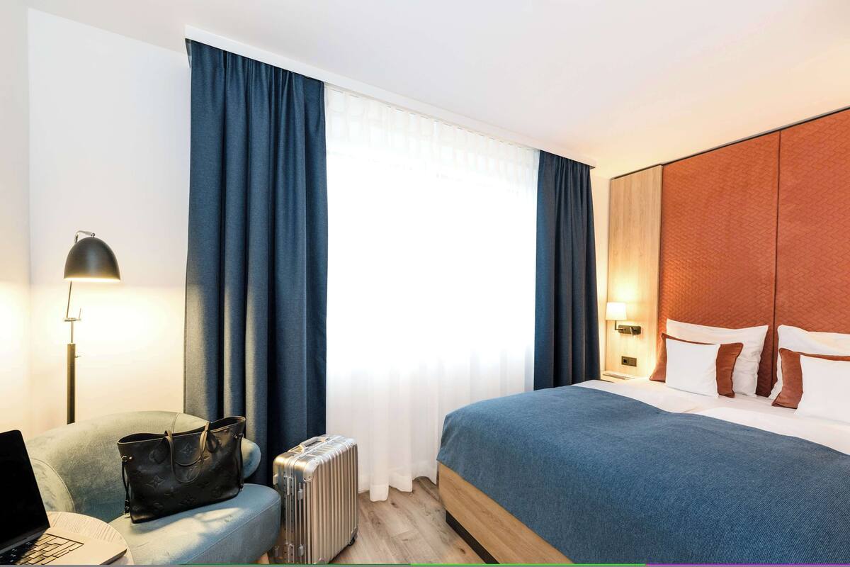 Een hotelkamer met een bed, een bureau met een laptop, een koffer en een handtas.