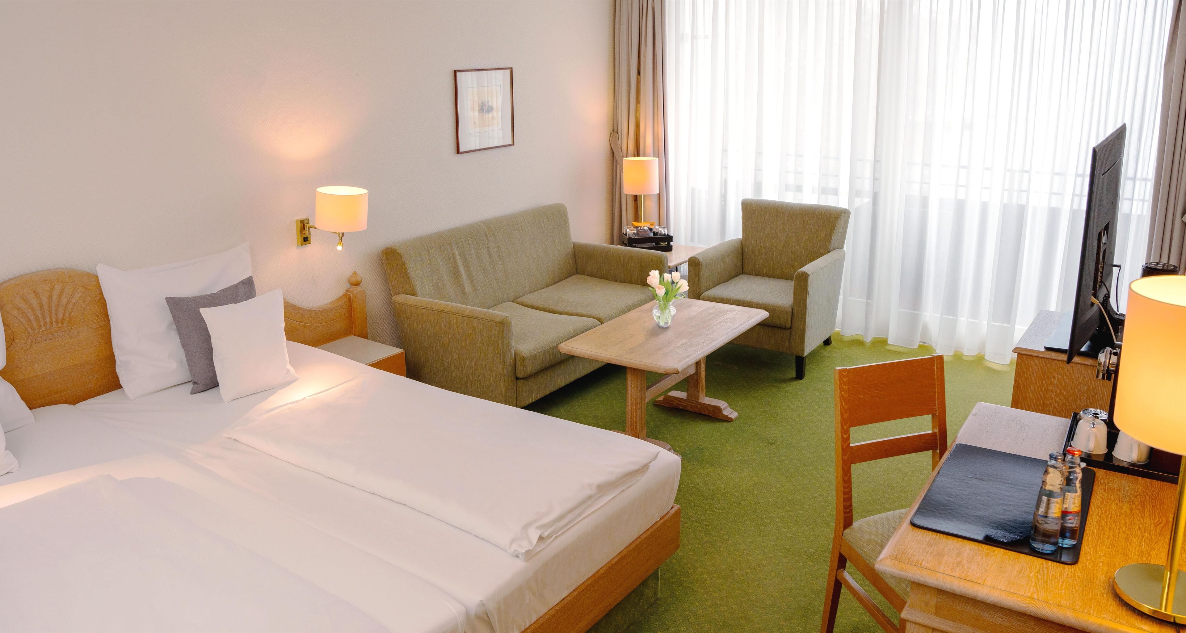 A hotel room with a bed, two armchairs, a small table, a desk with a chair, a TV, and a window with curtains.