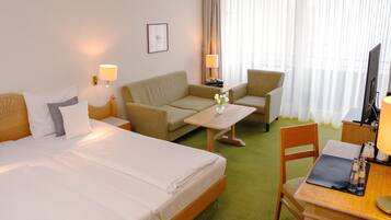 A hotel room with a bed, two armchairs, a small table, a desk with a chair, a TV, and a window with curtains.