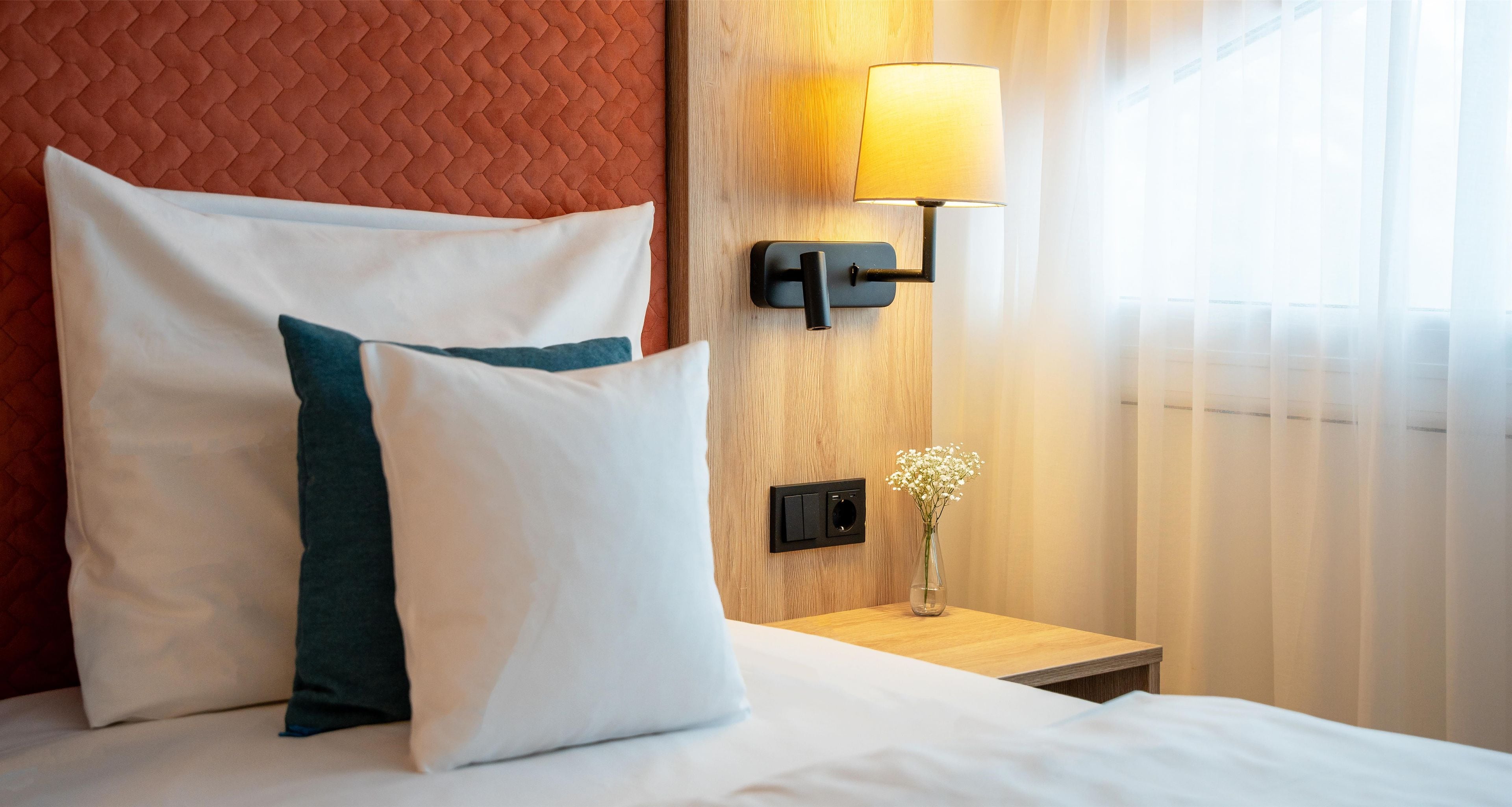 Ein ordentlich bezogenes Bett mit weißen und türkisfarbenen Kissen, einer Nachttischlampe und einem kleinen Blumenvasen auf einem Holznachttisch.