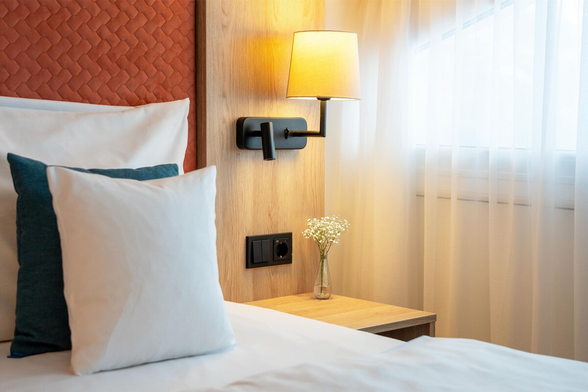 A hotel room with a bed, pillows, a nightstand, a lamp, a vase with flowers, and a wooden headboard.