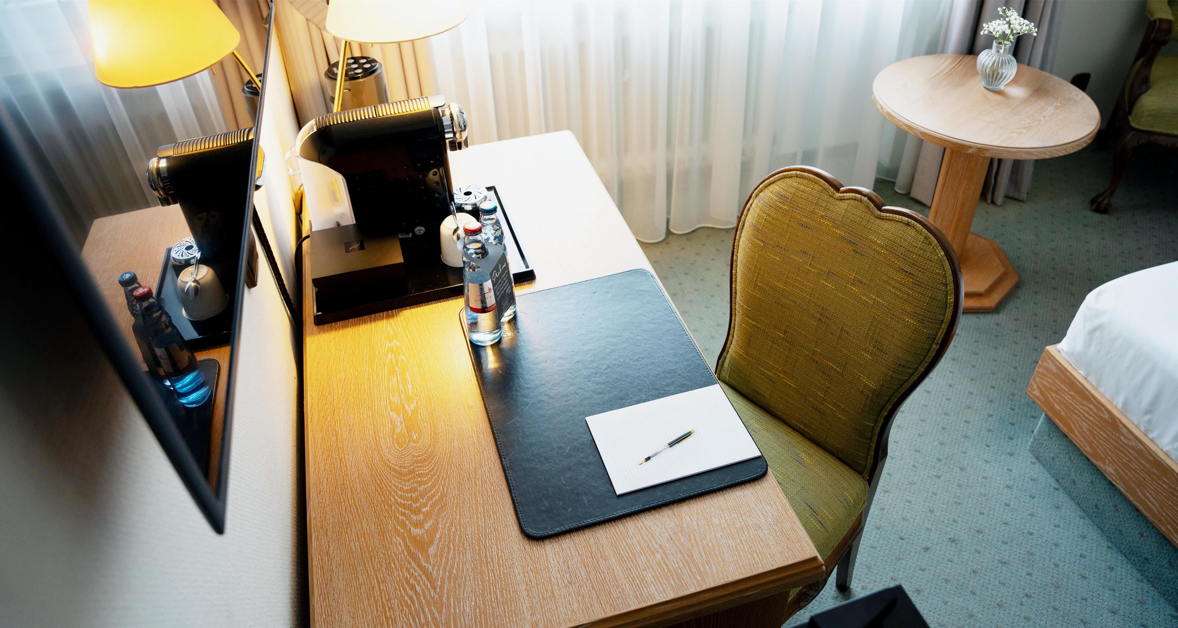 Une chambre d’hôtel avec un bureau en bois, un ordinateur portable, un stylo, une bouteille d’eau, une lampe et une chaise.