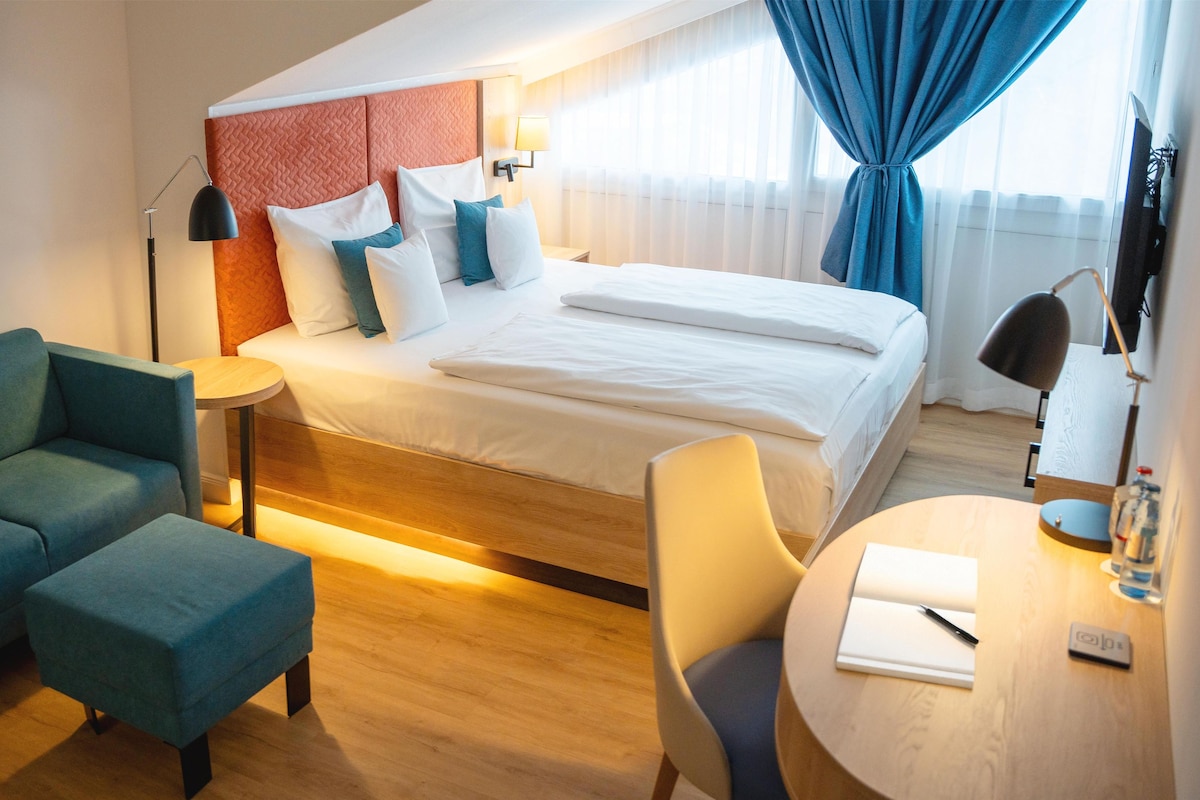 A modern hotel room with a bed, a sofa, a chair, a desk, and a lamp.