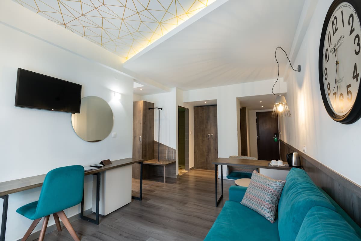 A modern hotel room with a large wall clock, a flat-screen TV, a sofa, a dining area, and a staircase leading to another room.