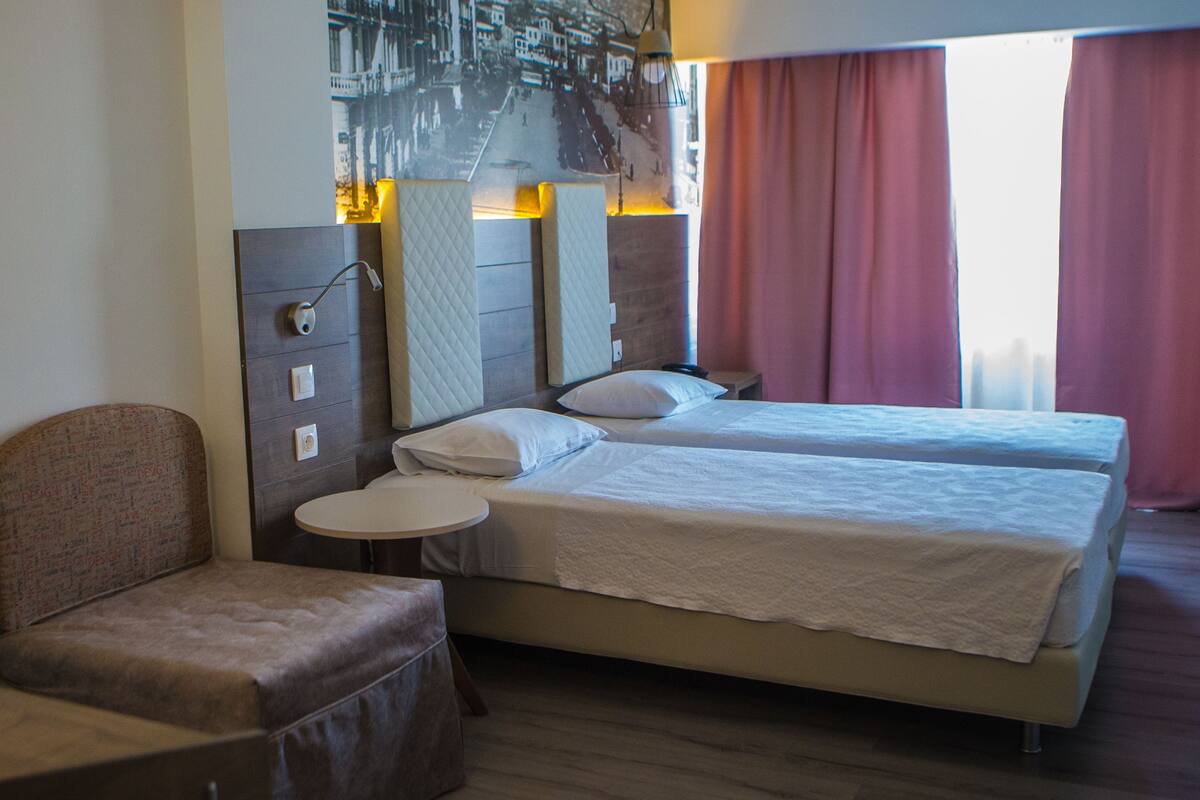 A hotel room with two beds, a small round table, a cushioned chair, and a wall-mounted TV.