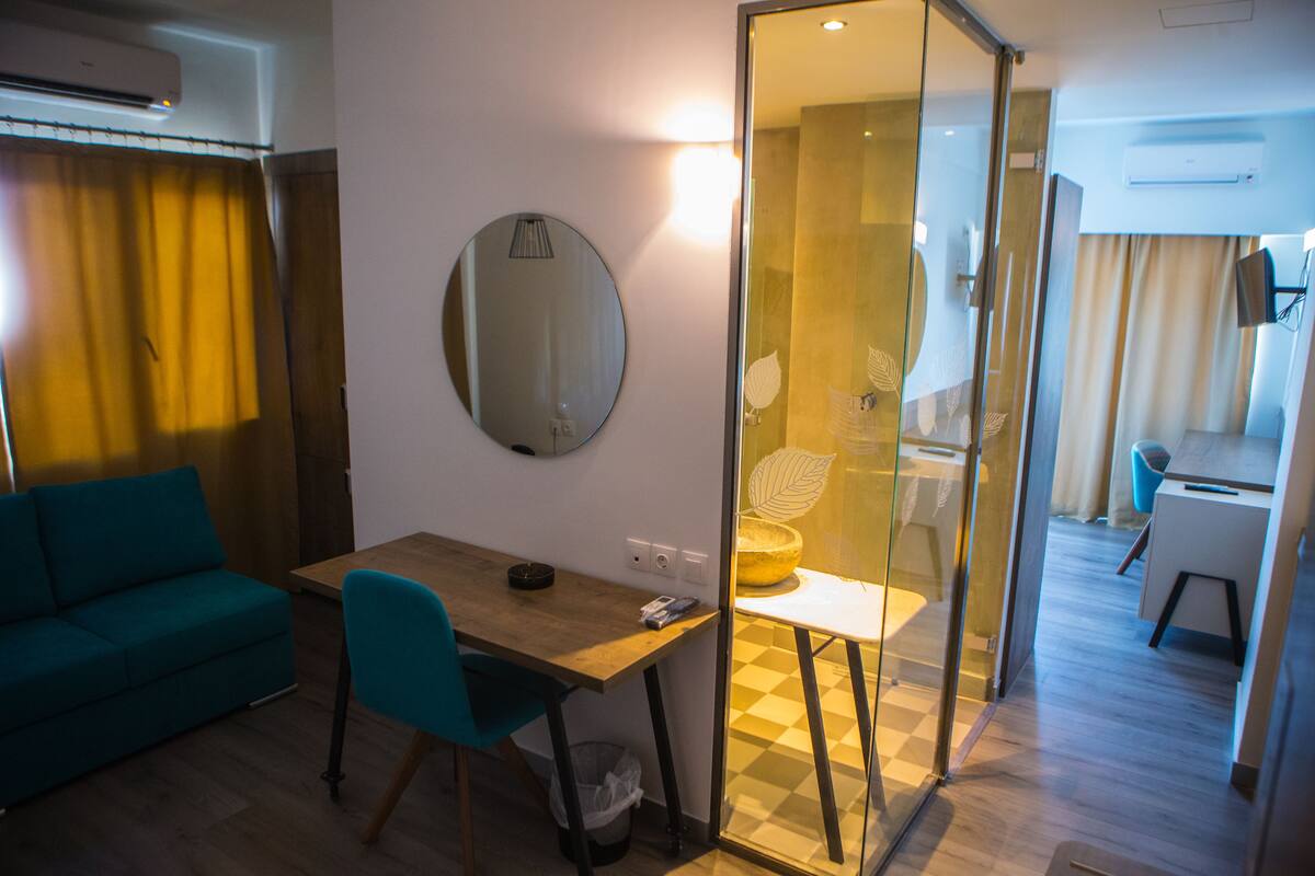 A modern hotel room with a wooden desk, a teal sofa, a round mirror, and a glass partition separating the living area from the dining space.