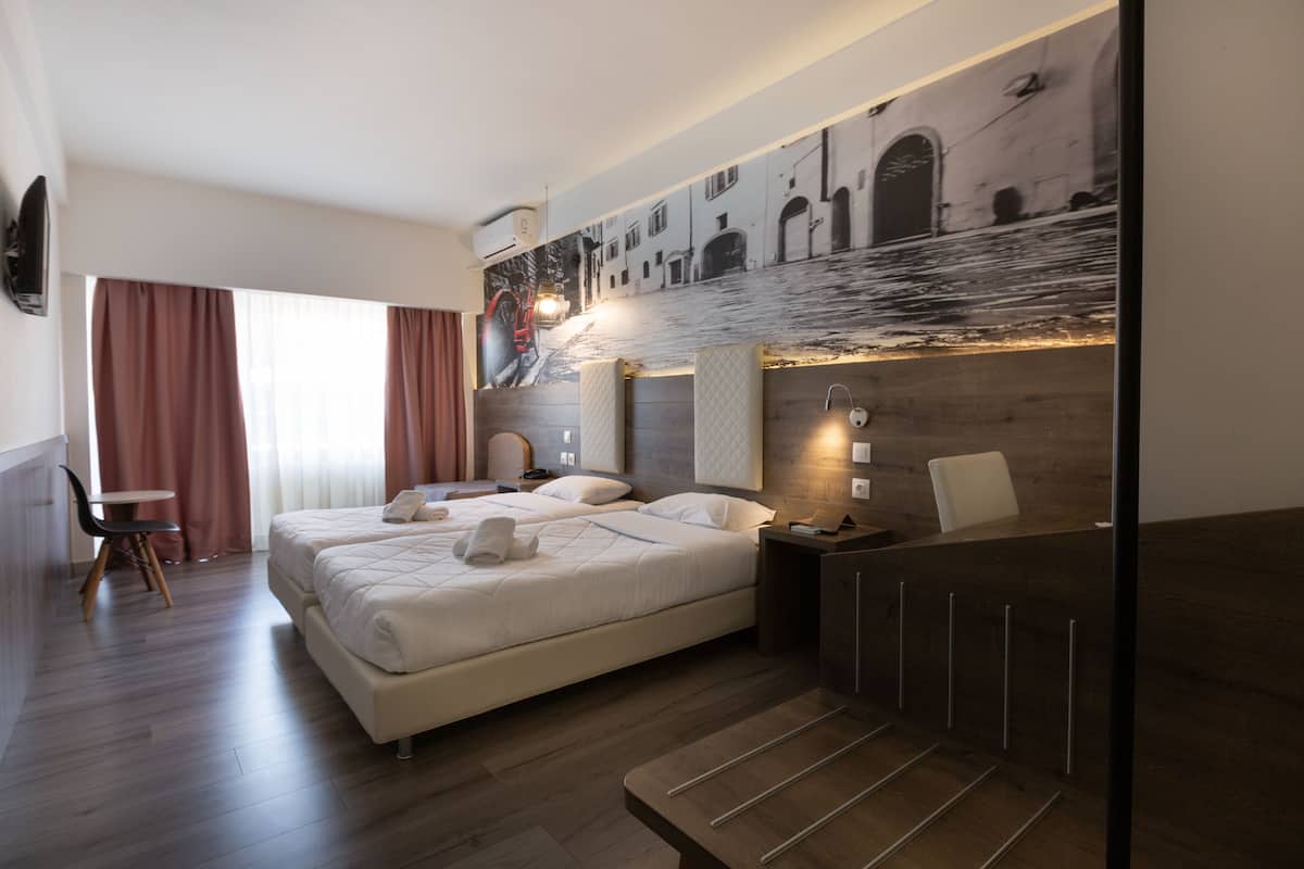A modern hotel room with a large bed, a desk, and a chair. There is a painting on the wall and a window with curtains.