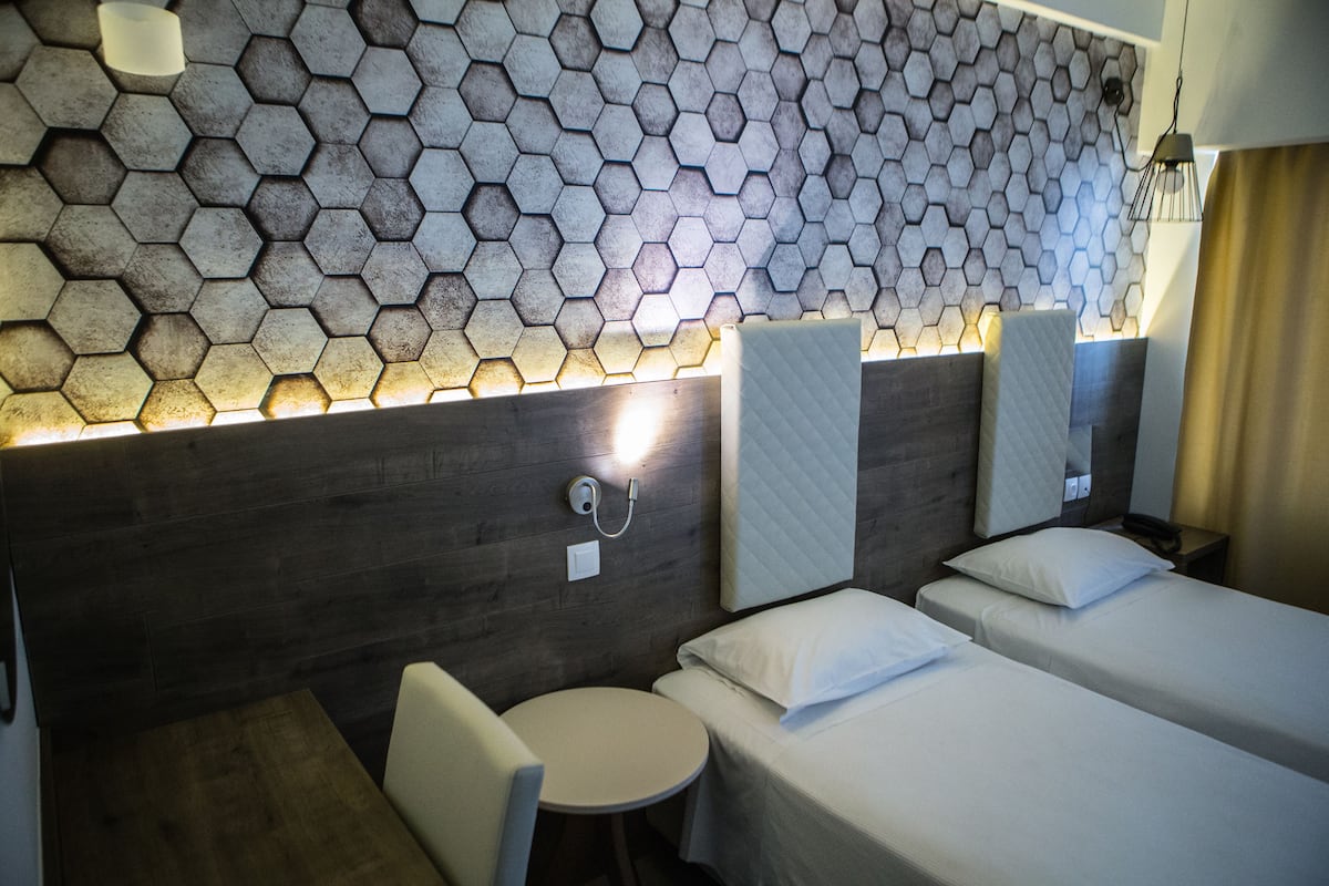A hotel room with a hexagonal patterned wall, a wooden headboard, two beds with white linens, a round table, and a chair.