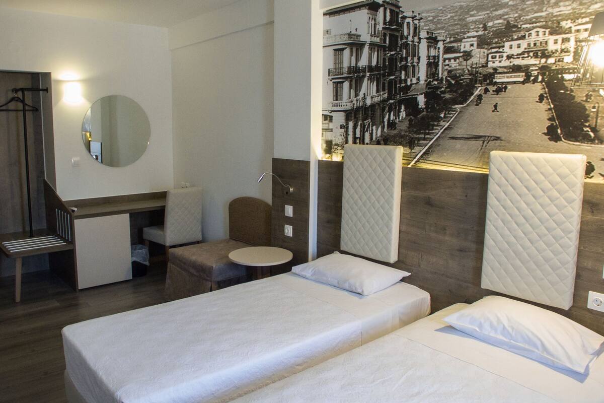 A hotel room with two beds, a desk, a chair, a round table, a mirror, and a view of a street scene.