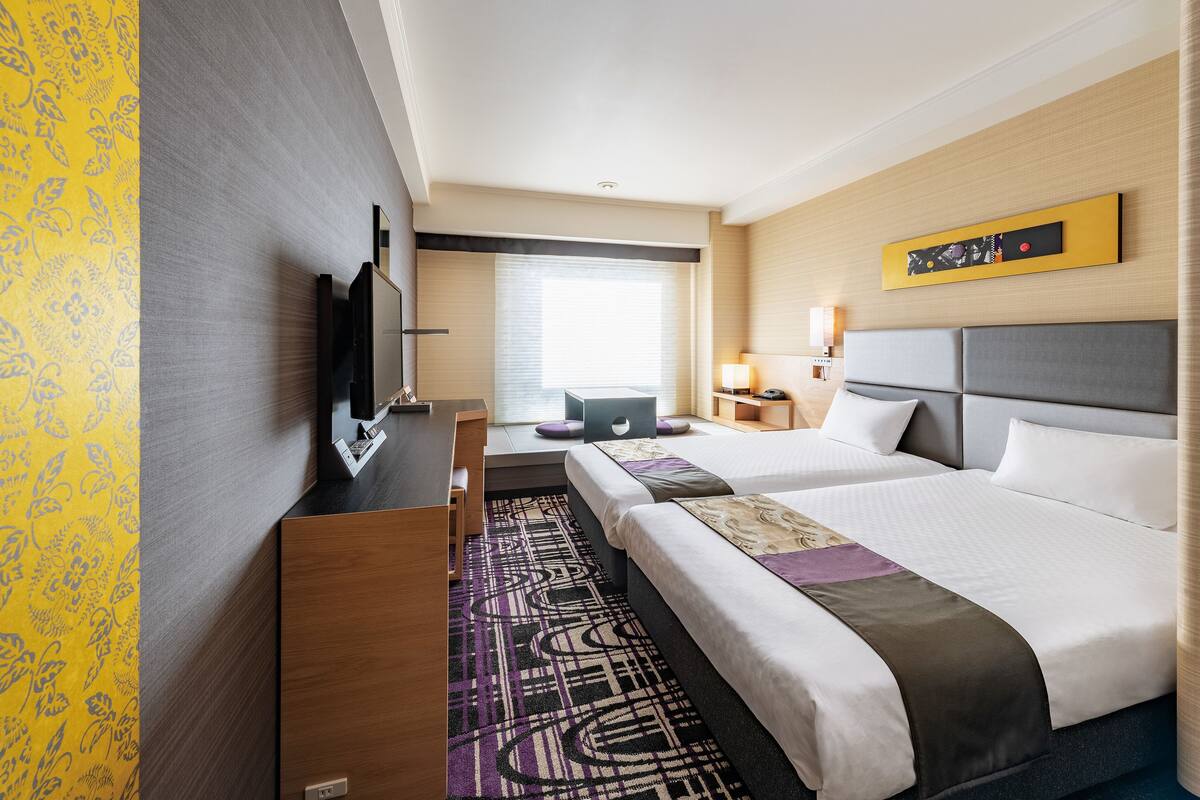 Ein Hotelzimmer mit zwei Betten, einem Schreibtisch, einem Flachbildfernseher und einem Fenster mit Jalousien.
