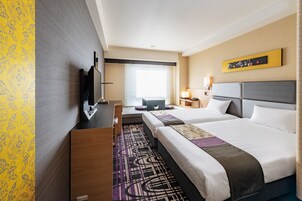 A hotel room with two beds, a desk, a flat-screen TV, and a window with blinds.