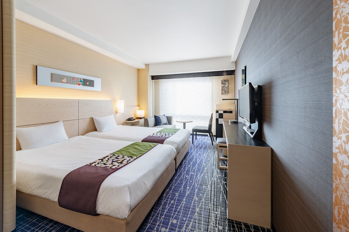 Ein Hotelzimmer mit zwei Betten, einem Schreibtisch, einem Fernseher und einem Fenster mit Jalousien.