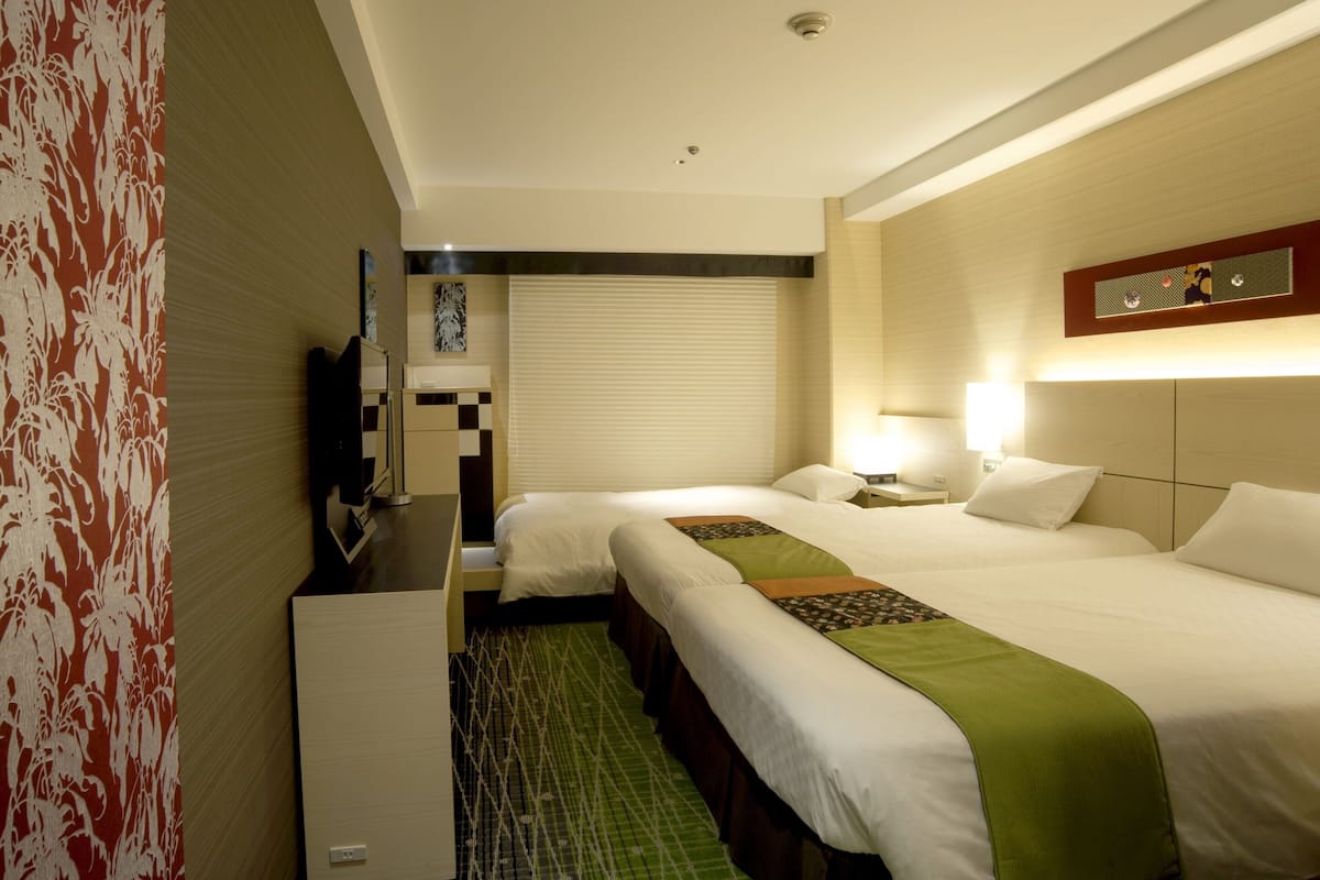 Ein Hotelzimmer mit zwei Betten, einem Schreibtisch, einem Fernseher und einem Fenster mit Jalousien.