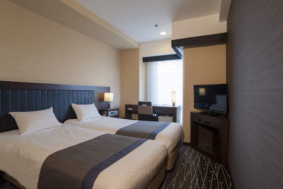 Ein Hotelzimmer mit zwei Betten, einem Schreibtisch, einem Fernseher und einem Fenster mit Jalousien.