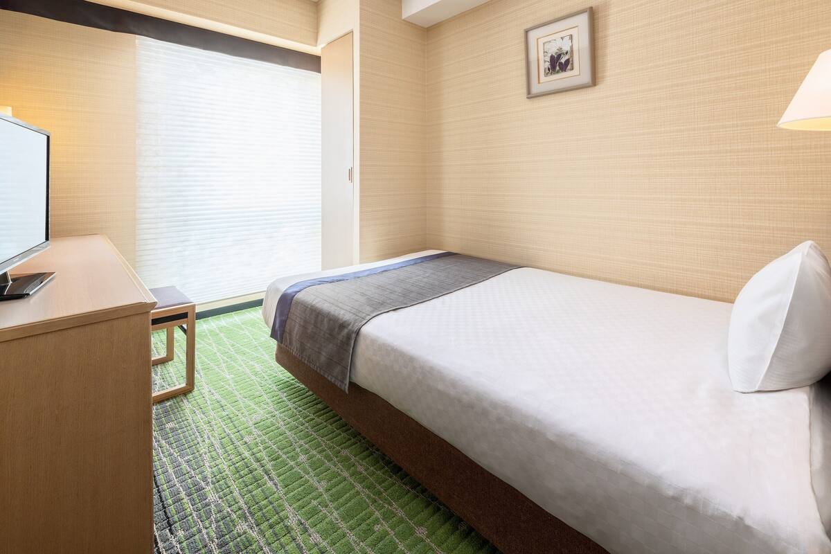 Ein Hotelzimmer mit Bett, Schreibtisch, Stuhl, Fenster mit Jalousien und einem gerahmten Bild an der Wand.