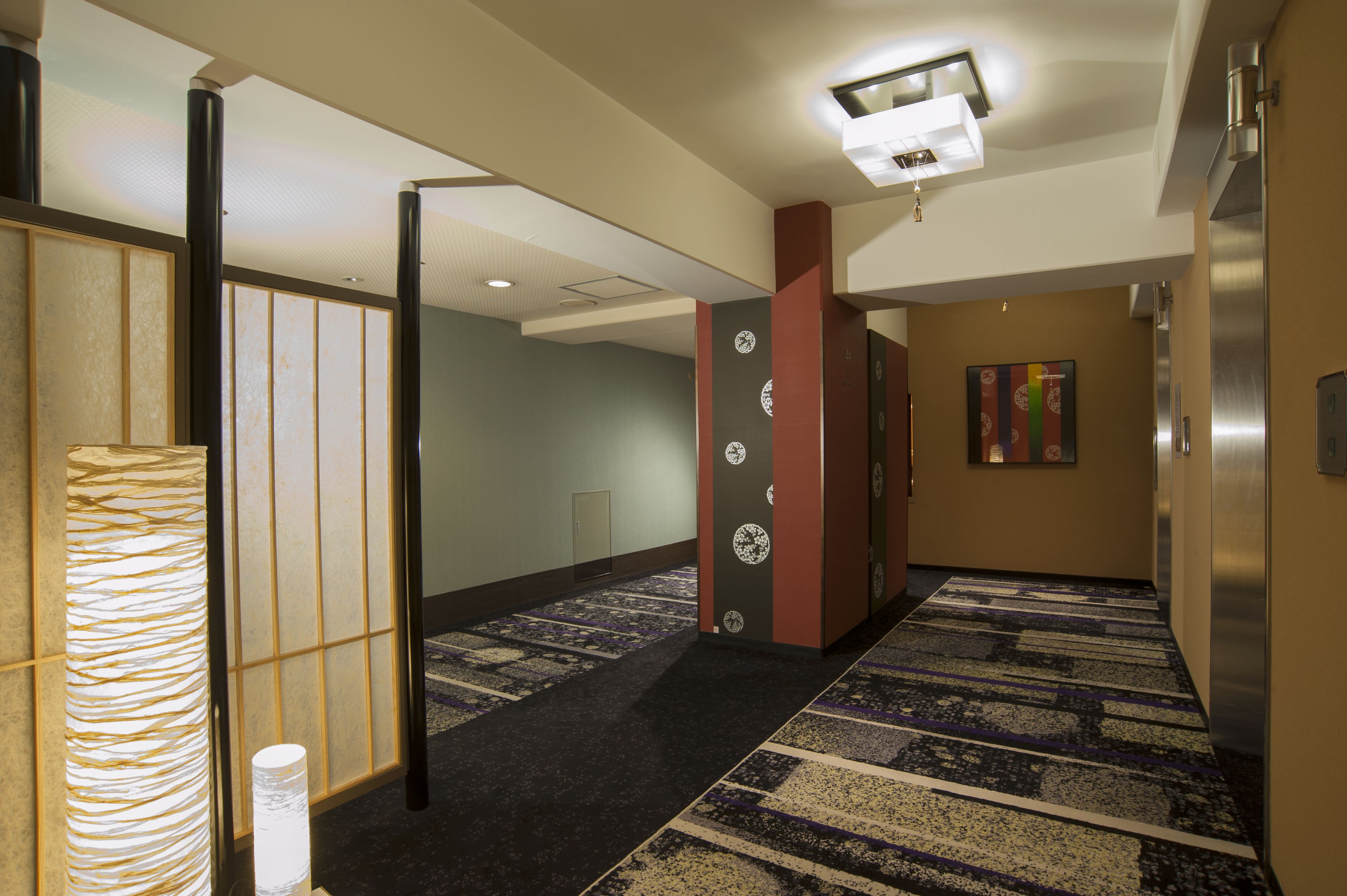 Un pasillo de hotel con alfombra estampada, una lámpara tradicional al estilo japonés y un moderno ascensor.