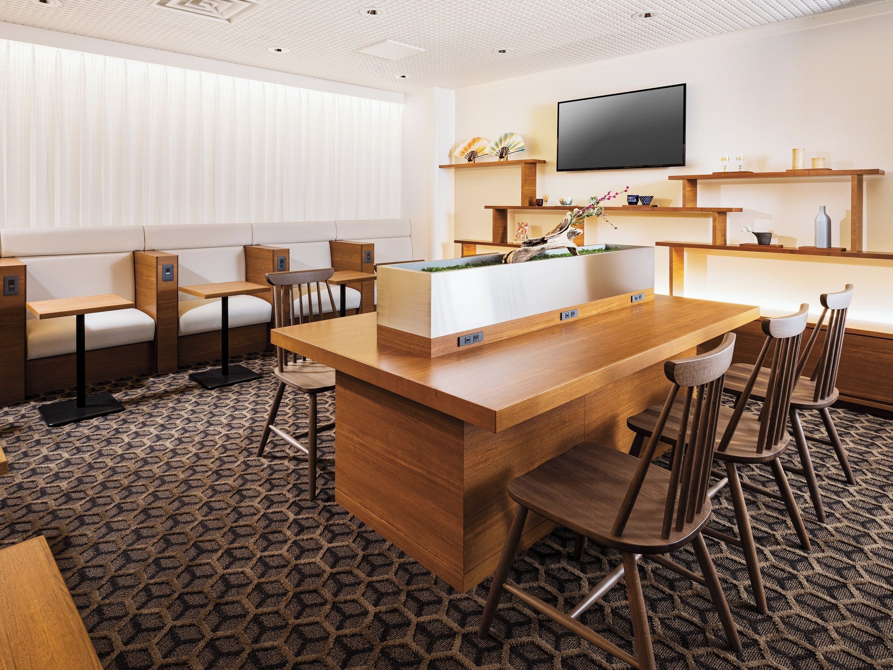 A modern hotel room with a central wooden bar, seating area, and a flat-screen TV mounted on the wall.