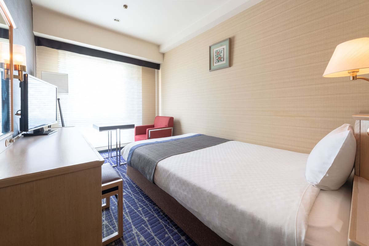 Ein Hotelzimmer mit Bett, Schreibtisch, Stuhl, kleinem Tisch und einem Fenster mit Jalousien.