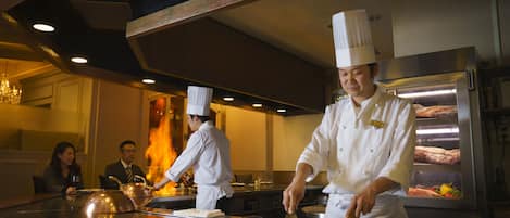 A professional kitchen with chefs preparing food, a stainless steel counter, and a large oven in the background.