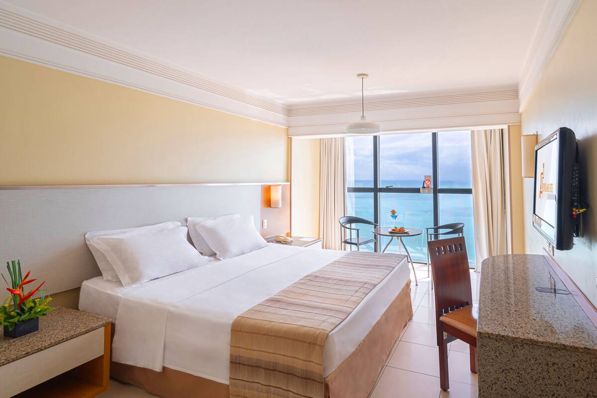 A hotel room with a large bed, a desk, a chair, a TV, and a view of the sea.