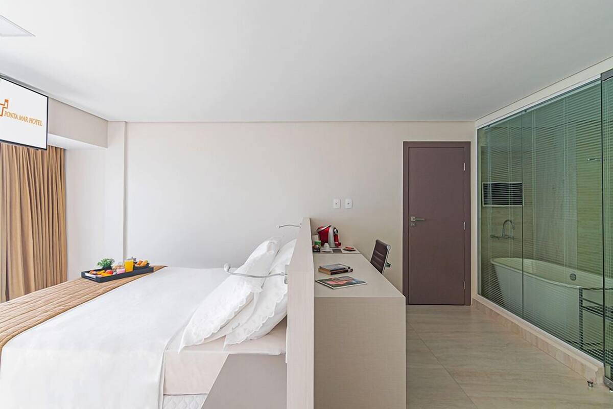 A modern hotel room with a bed, a small desk, a glass-enclosed shower, and a door.