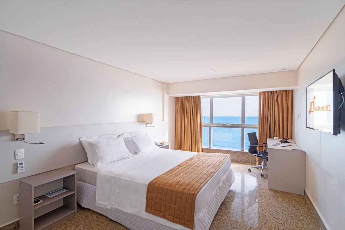A hotel room with a bed, desk, chair, and a large window offering a view of the sea.