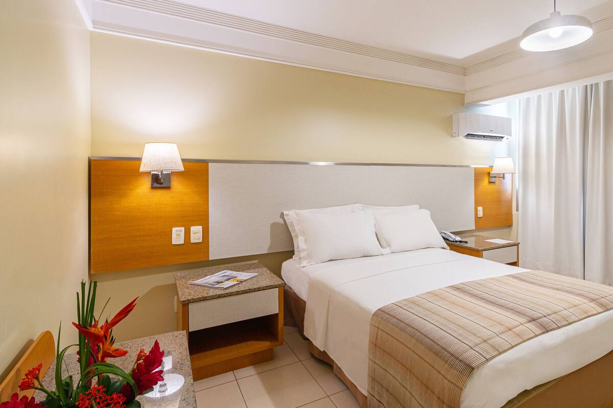 A hotel room with a bed, nightstands, a lamp, an air conditioner, and a window with curtains.