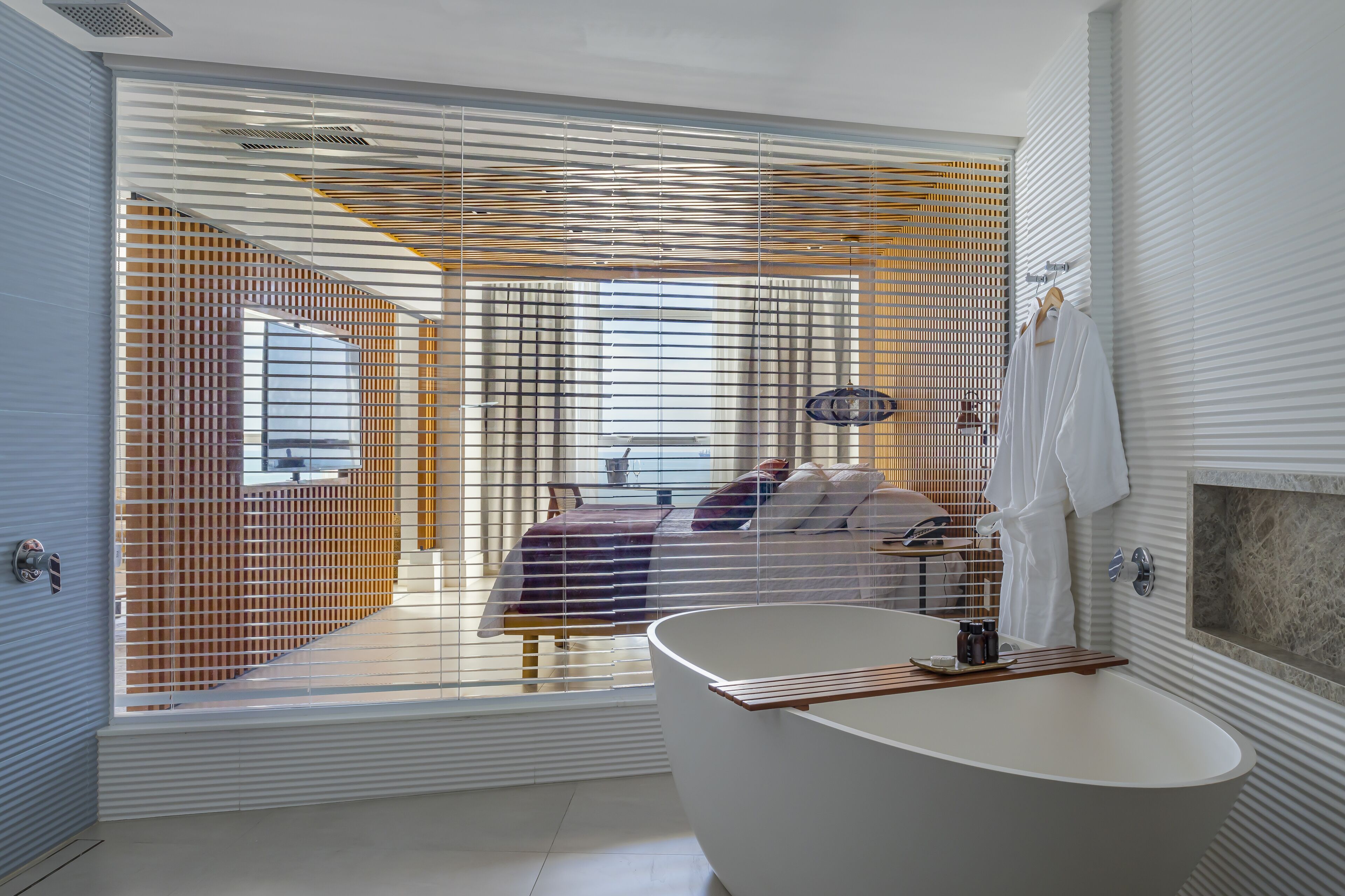 Un baño moderno con una bañera independiente, zona de ducha y un dormitorio visible con cama y persianas.