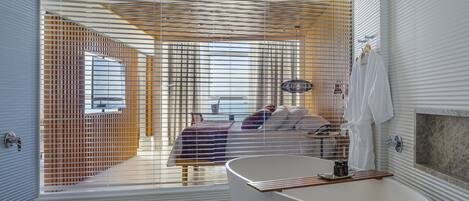Un baño moderno con una bañera independiente, zona de ducha y un dormitorio visible con cama y persianas.