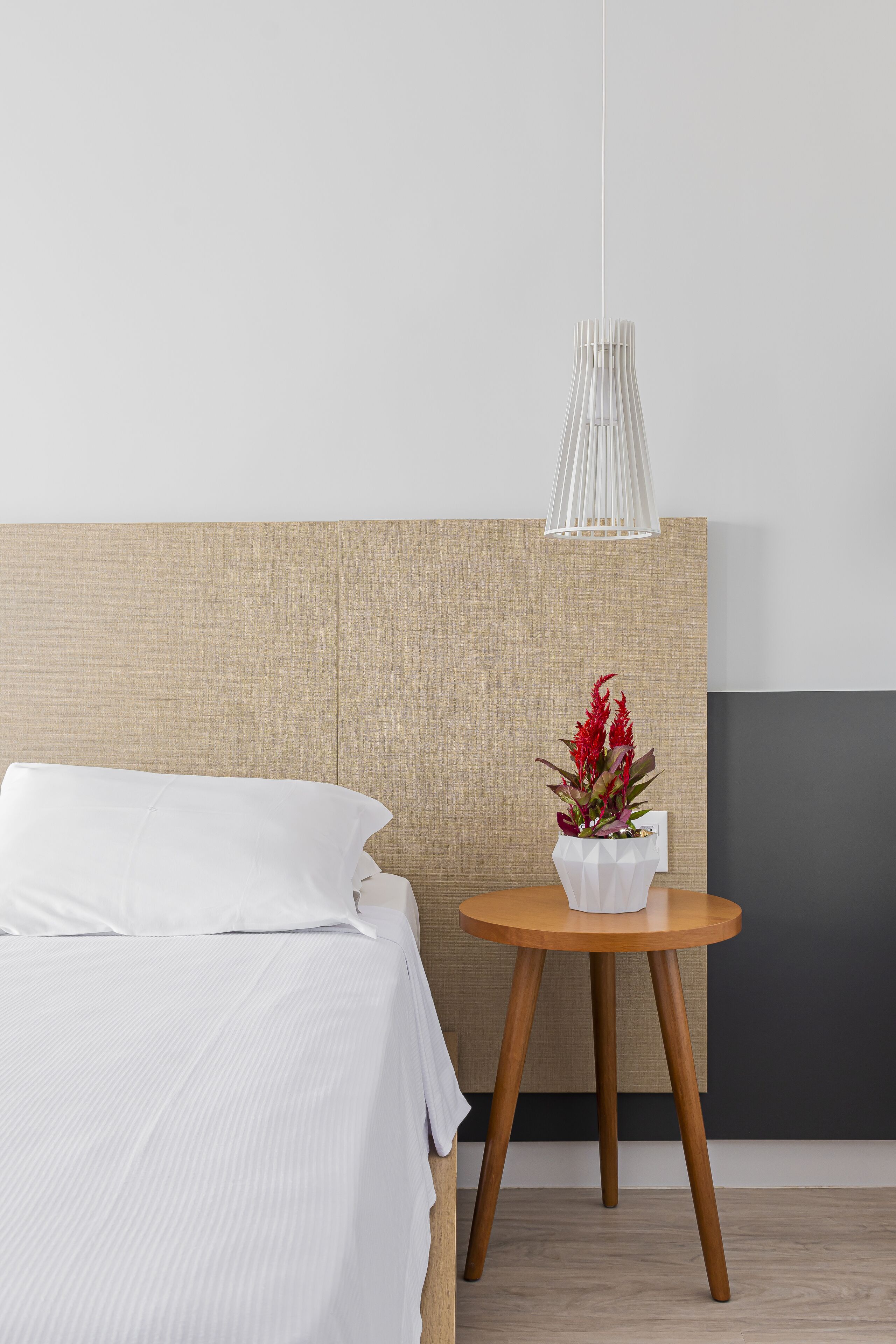 A neatly made bed with white linens, a round wooden side table with a white vase of red flowers, and a modern pendant light.