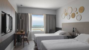 Habitación de hotel con dos camas, televisión, escritorio y silla, y vista al mar.