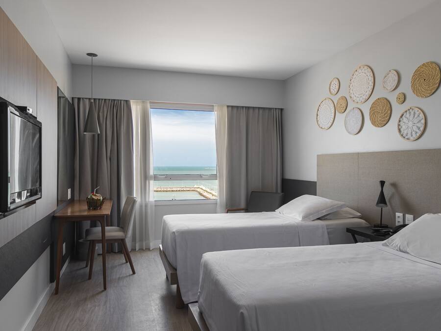 Habitación de hotel con dos camas, televisión, escritorio y silla, y vista al mar.