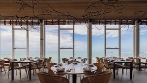 Restaurante com grandes janelas que oferecem vista para o mar. Mesas montadas com pratos, copos e talheres.