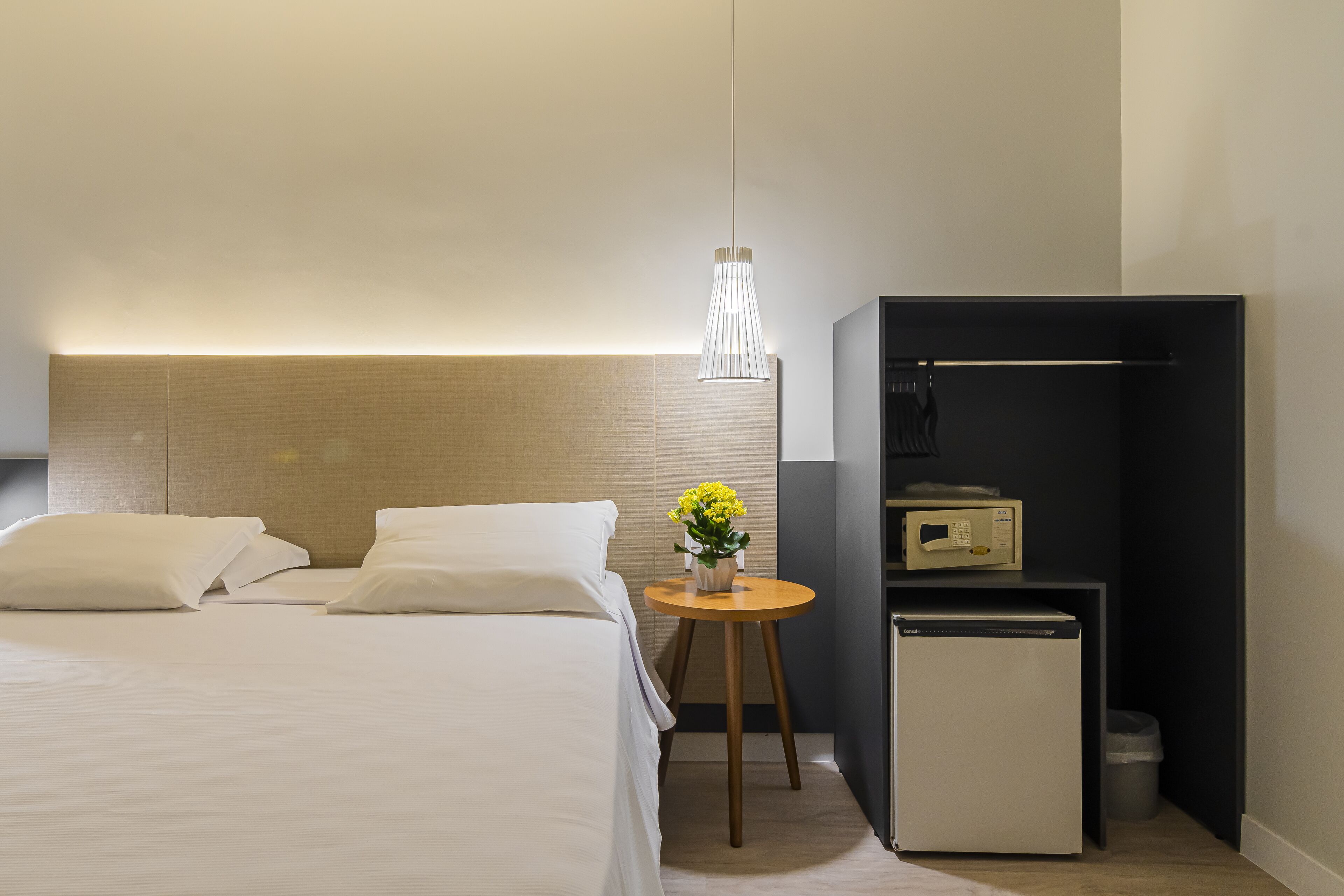 Une chambre d’hôtel moderne avec un lit soigneusement fait, une petite table ornée d’un vase de fleurs, un mini-réfrigérateur et une armoire intégrée.