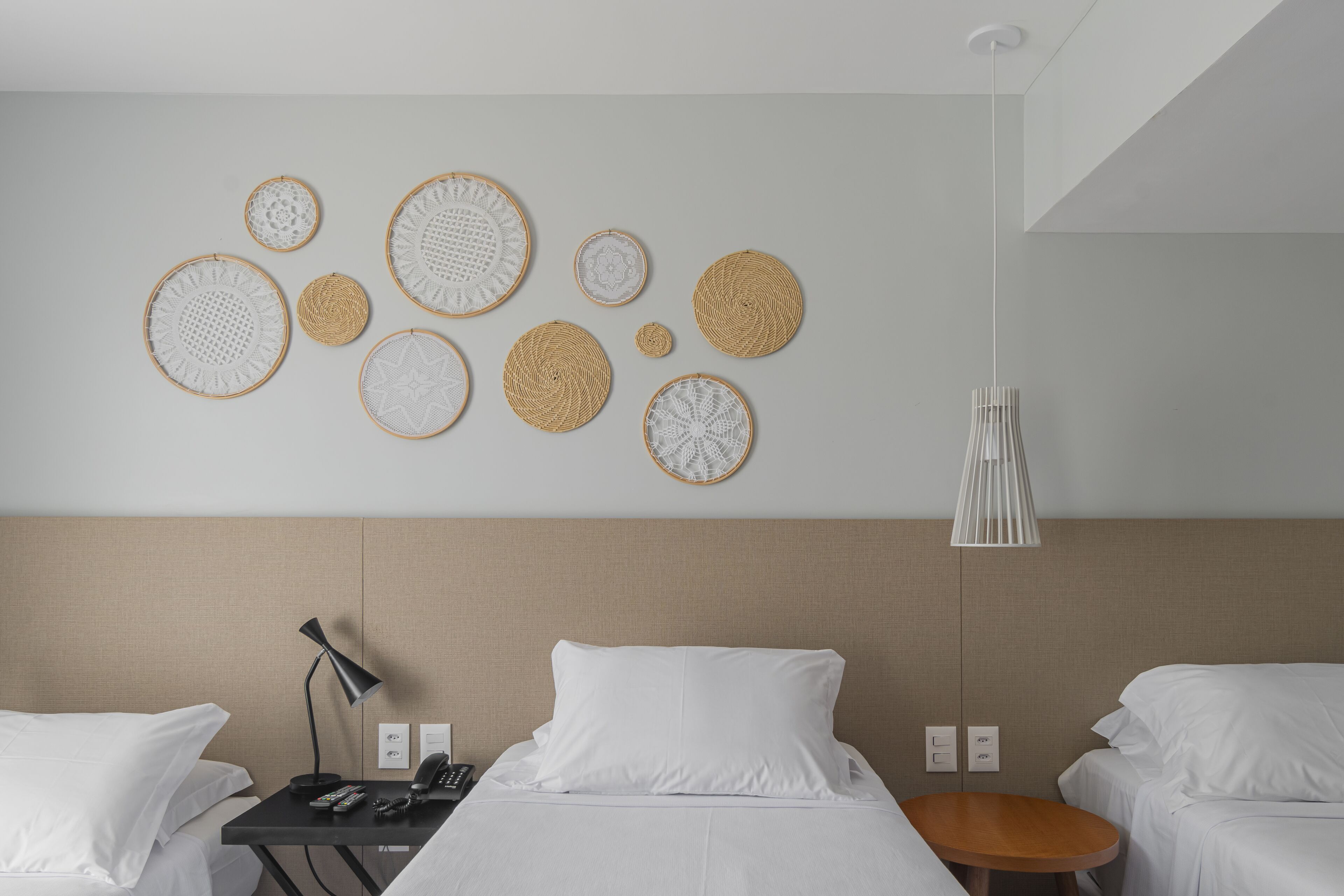 A hotel room with two beds, a bedside table with a lamp, a wall-mounted phone, and a hanging light fixture.