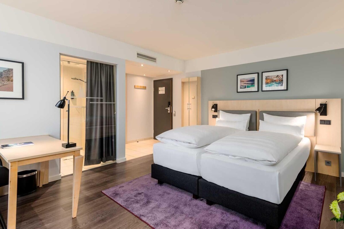 Ein modernes Hotelzimmer mit einem großen Bett, einem Schreibtisch und einem Sessel. An der Wand hängen zwei gerahmte Bilder und ein Fenster mit Vorhängen.
