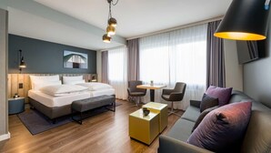 A modern hotel room with a large bed, a sofa, a coffee table, and a desk. There are two chairs, a TV, and a painting on the wall.