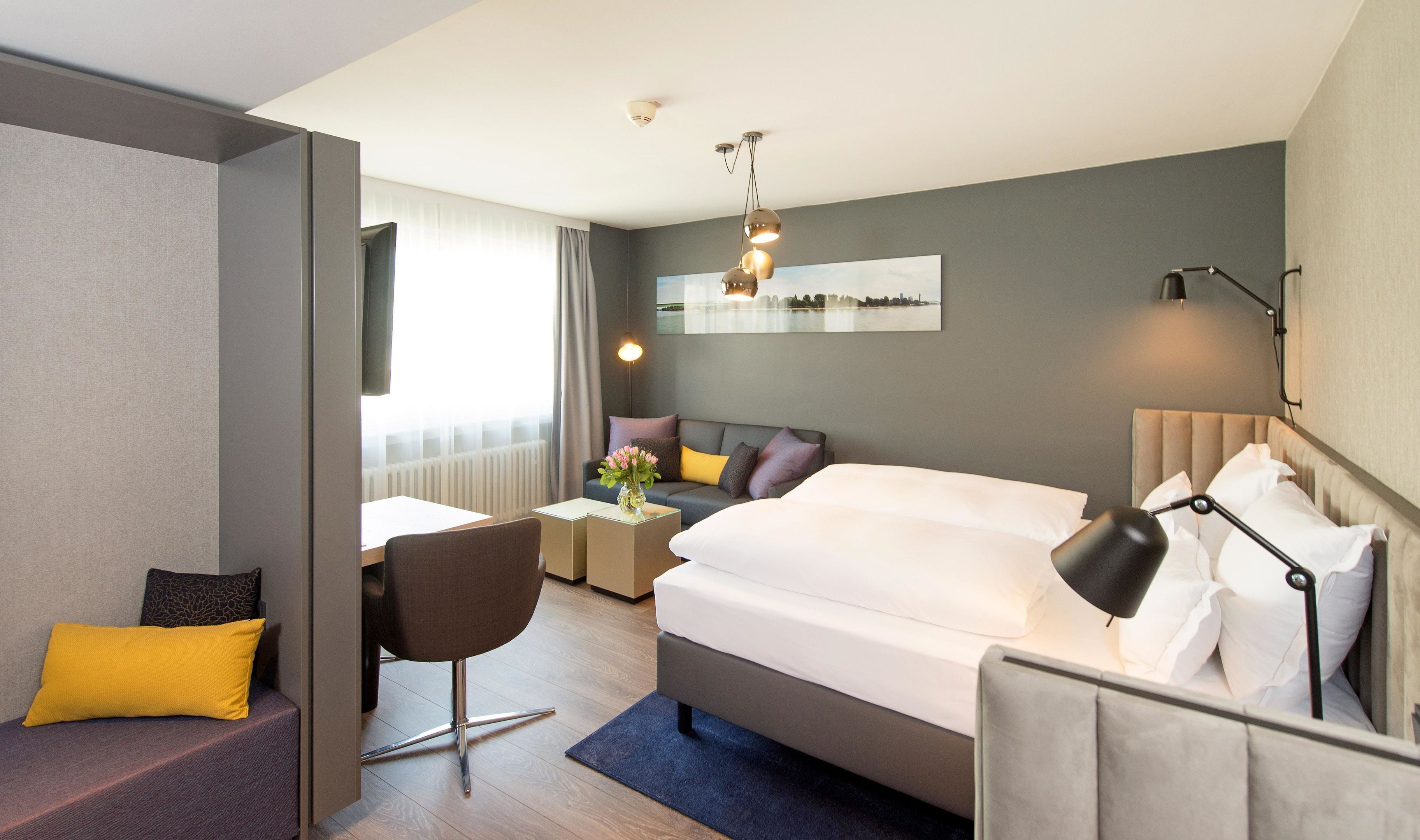 A modern hotel room with a large bed, a desk, a chair, and a sofa. There is a window with curtains, a mirror on the wall, and a ceiling light fixture.