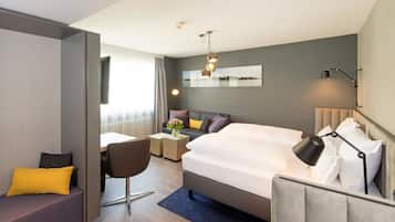 A modern hotel room with a large bed, a desk, a chair, and a sofa. There is a window with curtains, a mirror on the wall, and a ceiling light fixture.
