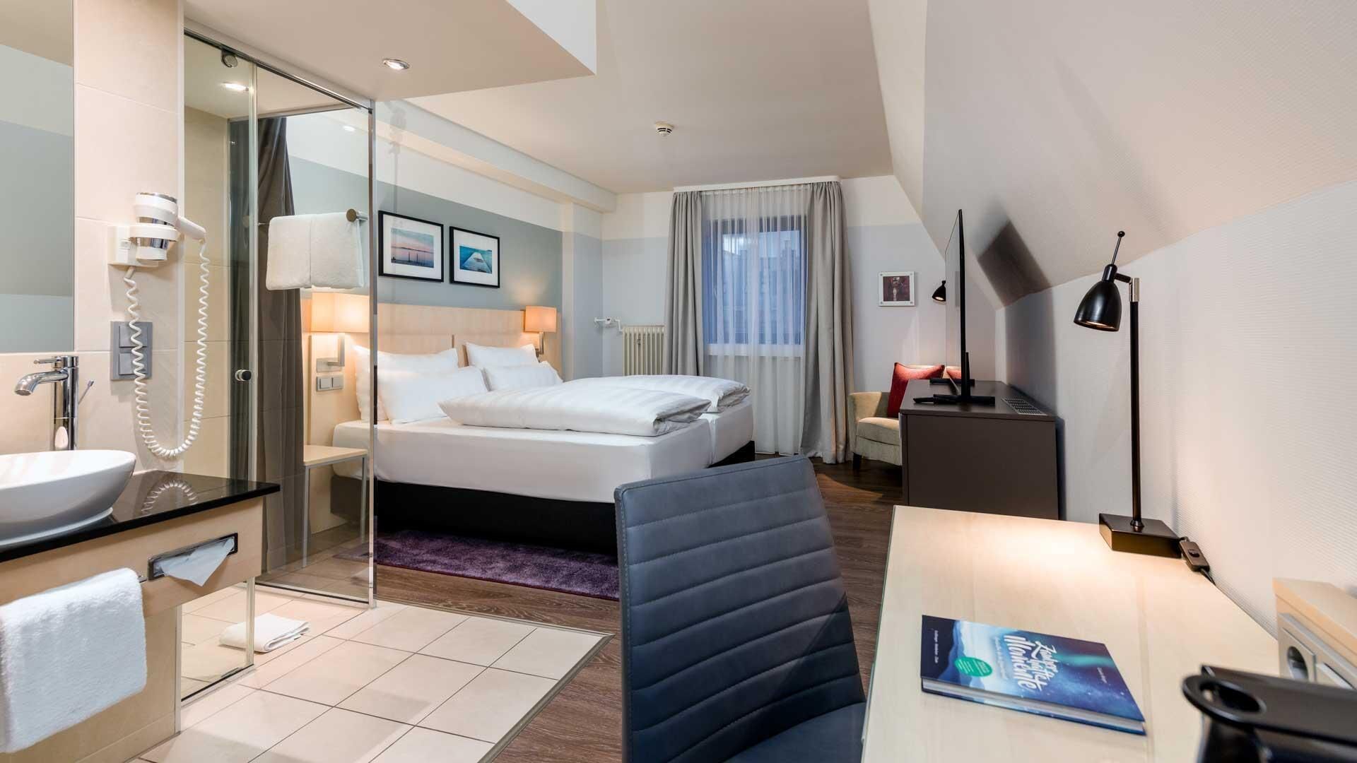 Een hotelkamer met een bed, nachtkastje, bureau, stoel, en een badkamer met een wastafel en spiegel.