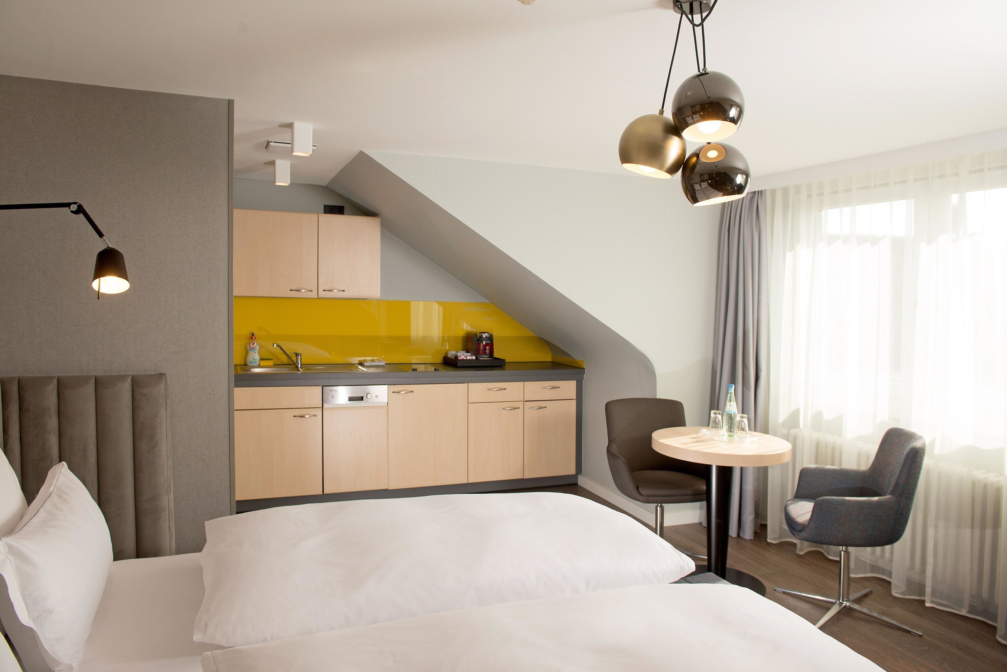 A modern hotel room with a bed, a desk, a chair, a small table, a kitchen area, and a dining area.