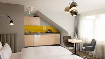 A modern hotel room with a bed, a desk, a chair, a small table, a kitchen area, and a dining area.