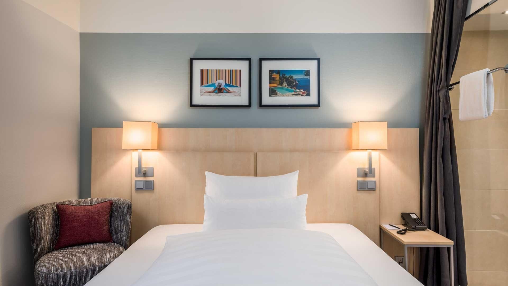 A hotel room with a bed, two wall-mounted lamps, a chair, a small table with a phone, and two framed pictures on the wall.
