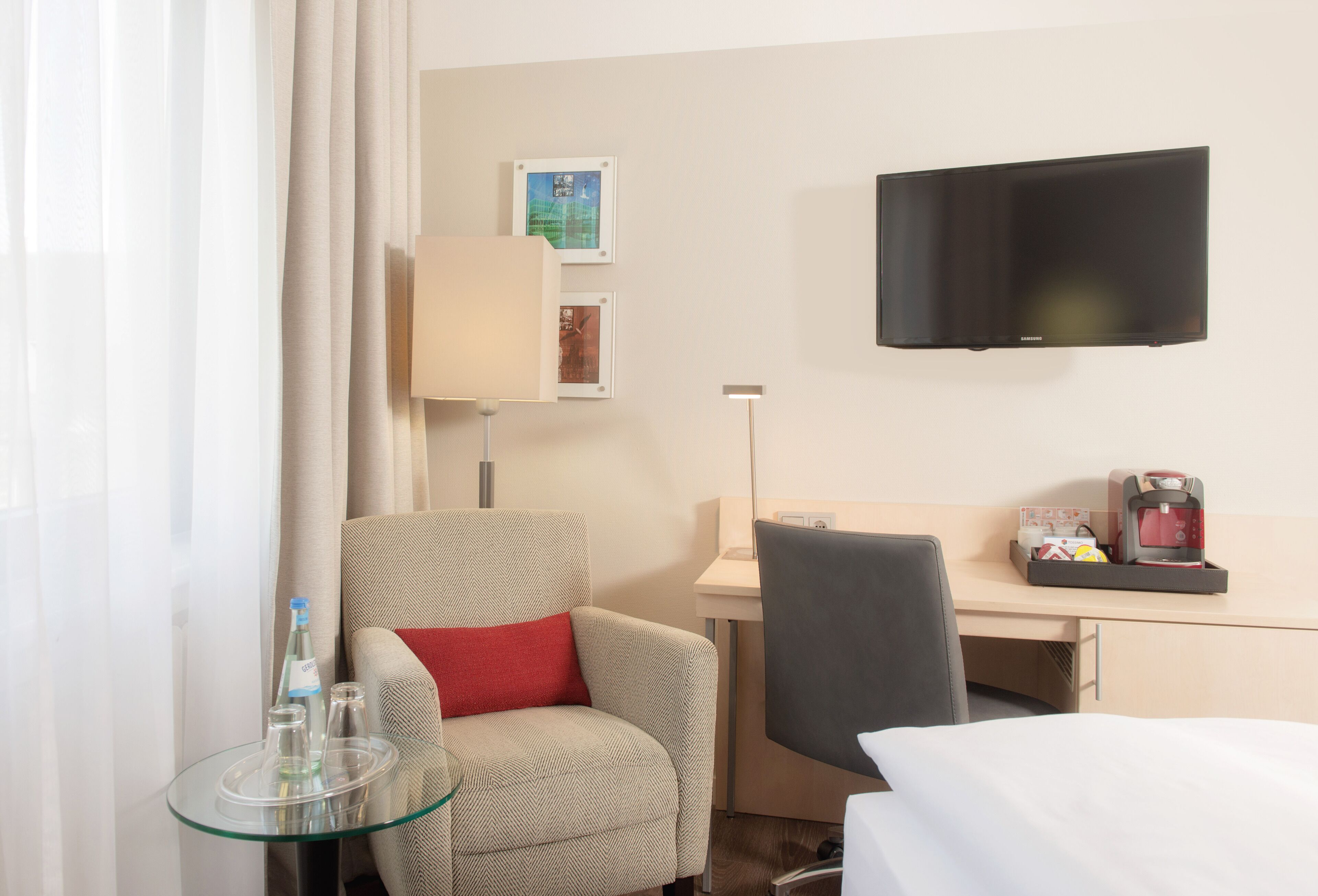 A hotel room with a bed, a desk, a chair, a TV, and a small table with a lamp and glasses.