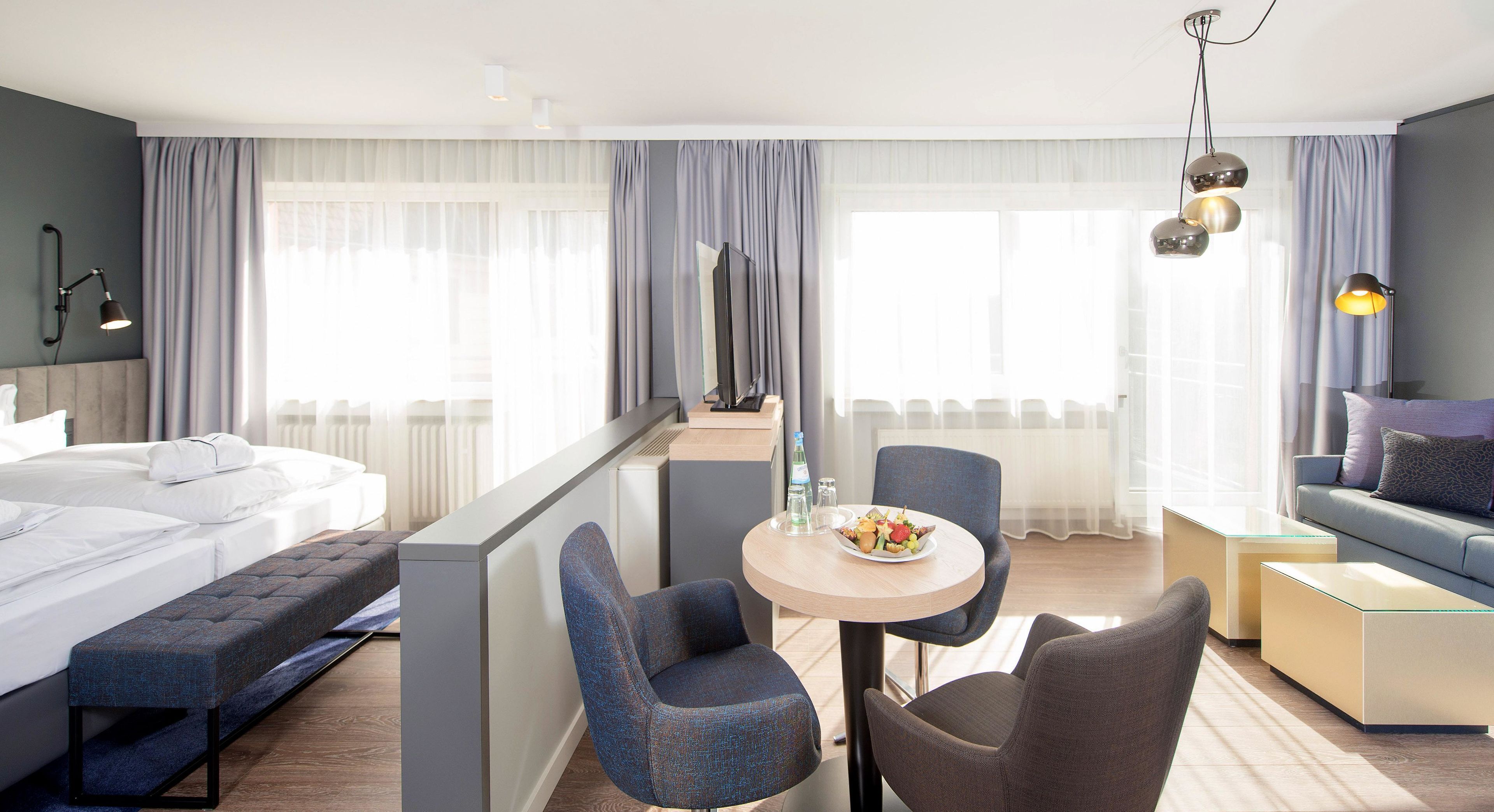 A modern hotel room with a bed, a dining area with two chairs, a sofa, and a TV.