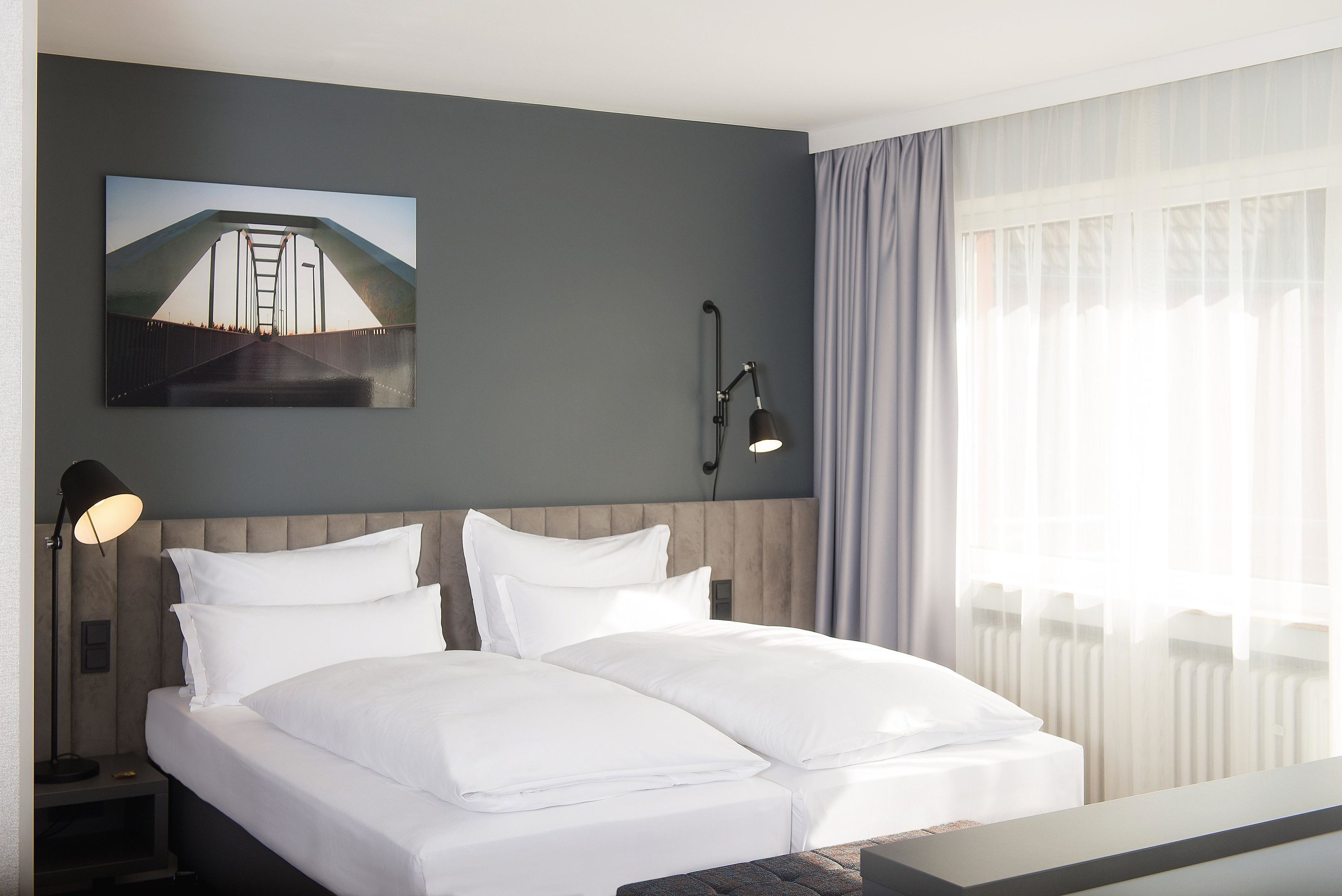 A hotel room with a bed, a nightstand, a lamp, a painting, and a window with curtains.