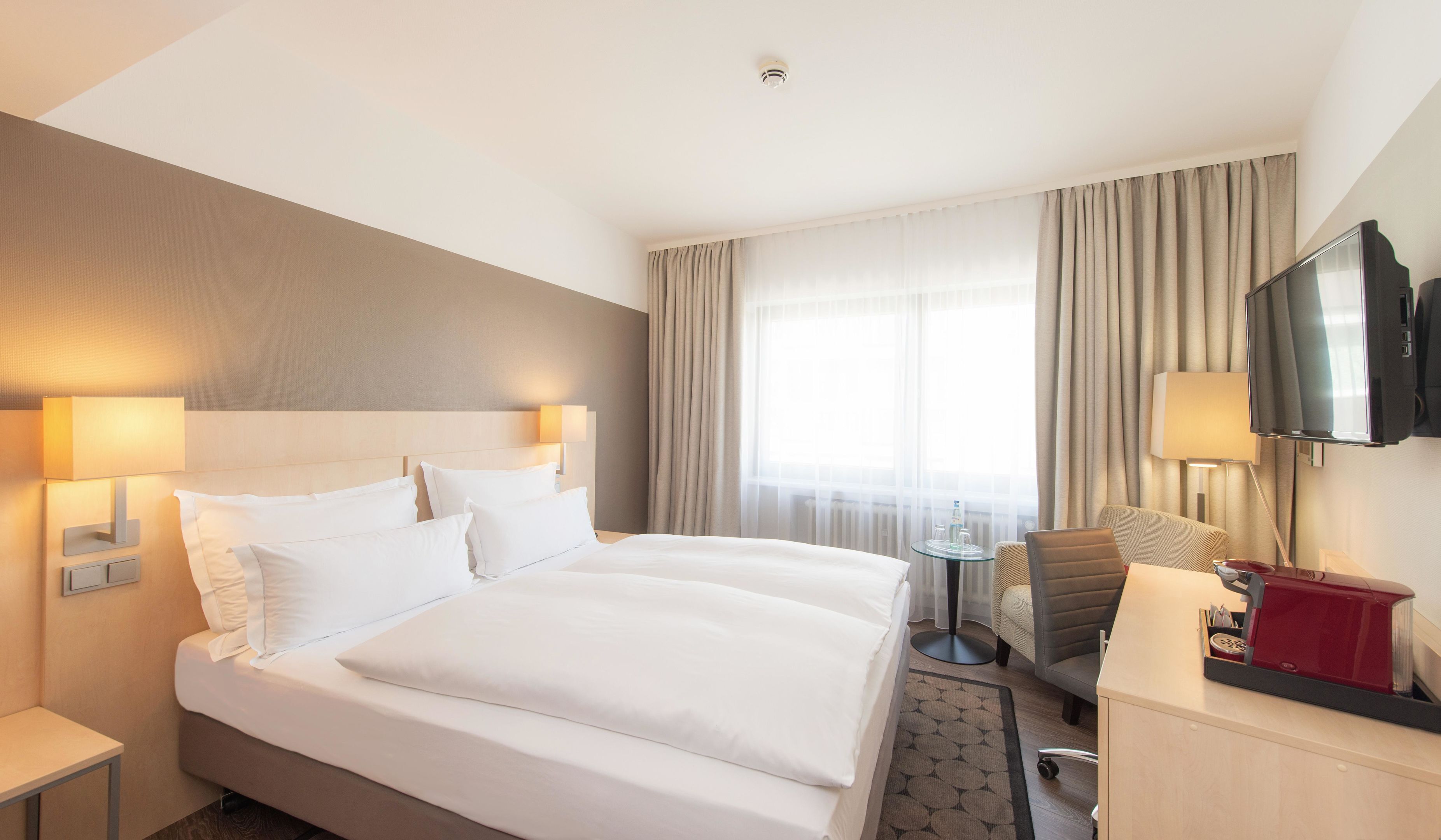 A hotel room with a large bed, two bedside lamps, a TV, a desk with a chair, and a coffee machine.