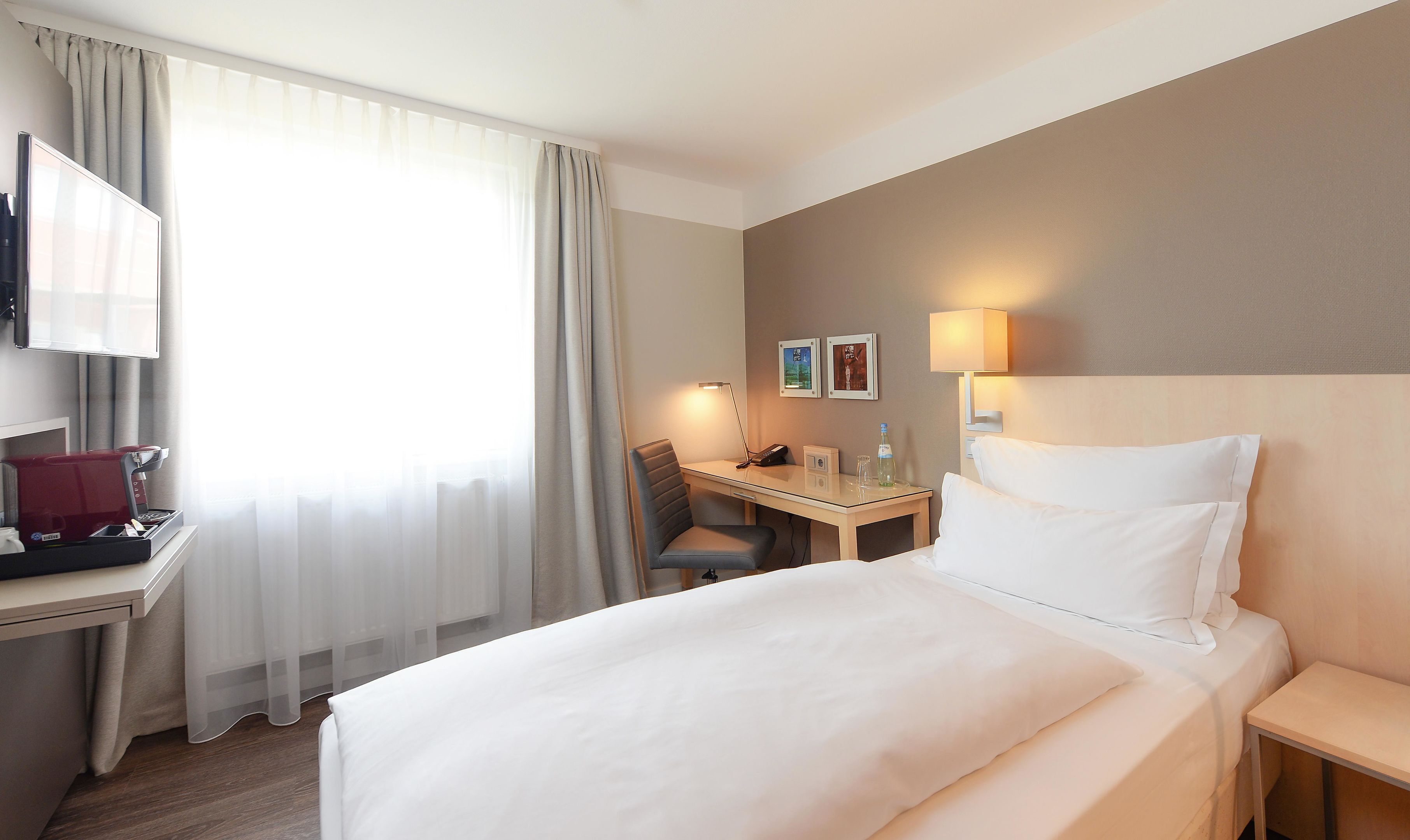 A hotel room with a bed, a desk with a computer, a chair, a coffee machine, and a TV.
